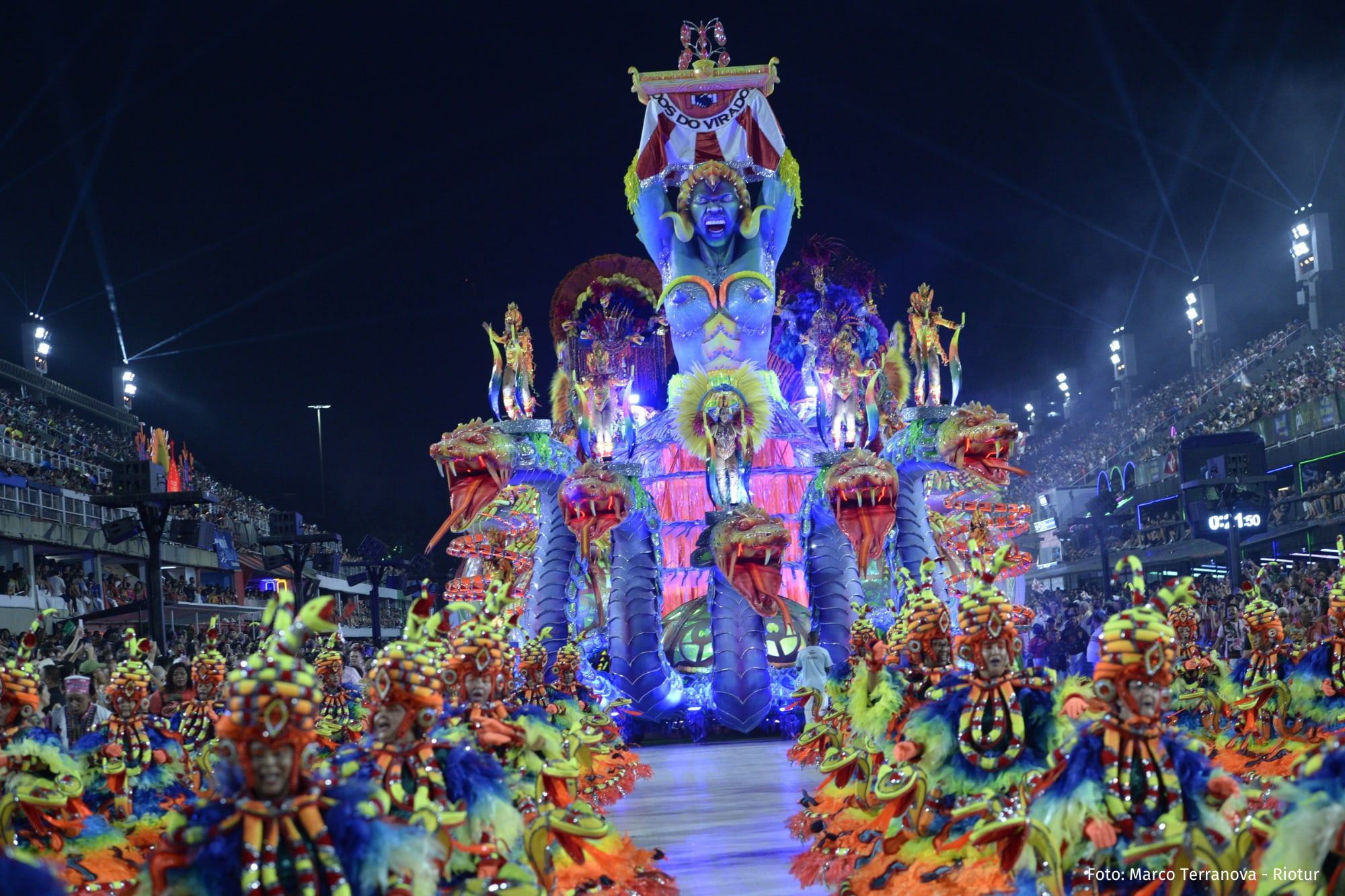 A towering illuminated float surrounded by costumed dancers at the Sambadrome at night